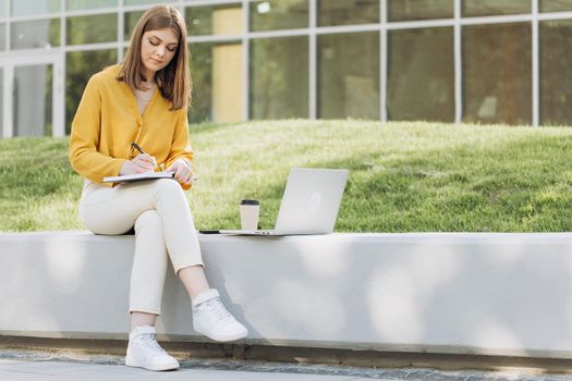 Student girl watching lesson online and studying outside home. Young woman taking notes while looking at computer screen following professor on video call. Girl student studying sitting outside.