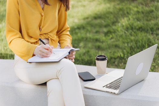 Student girl watching lesson online and studying outside home. Young woman taking notes while looking at computer screen following professor on video call. Girl student studying sitting outside.