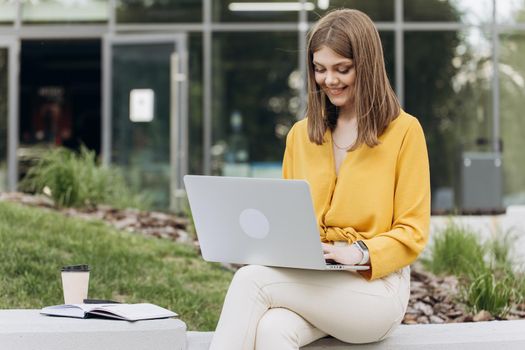 Attractive young female using laptop while sitting on bench outdoors in city. Pretty girl student preparing for classes in nature. Lady corporate start up office employee working outdoor