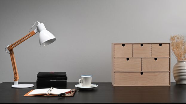 Lamp, small wooden drawer, books and coffee cup on black table.