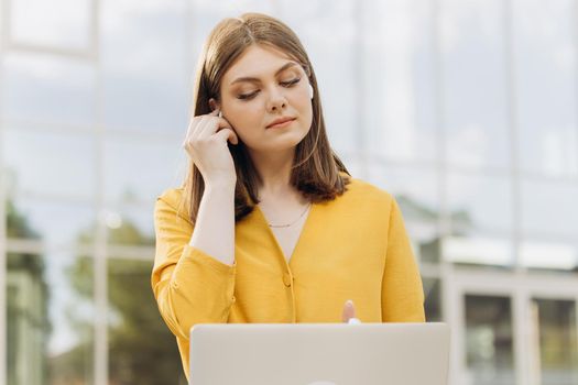 Joyful attractive woman wearing earphones, looking at laptop computer while sitting at outdoor. Female manager typing on laptop keyboard. Portrait of smiling business woman looking at laptop screen.