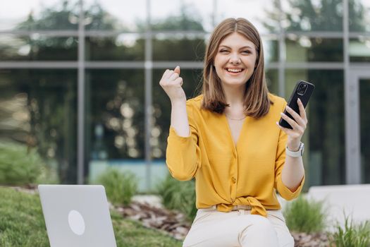 Surprised lady celebrating victory on smartphone outdoor. Portrait of happy business caucasian woman enjoy success on mobile phone. Joyful girl reading good news on phone.