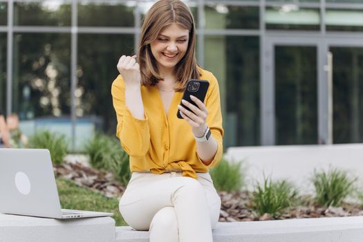 Portrait of happy business caucasian woman enjoy success on mobile phone. Joyful girl reading good news on phone. Surprised lady celebrating victory on smartphone outdoor.