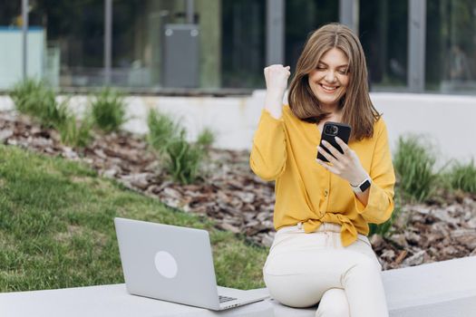 Joyful girl reading good news on phone. Surprised lady celebrating victory on smartphone outdoor. Portrait of happy business caucasian woman enjoy success on mobile phone.