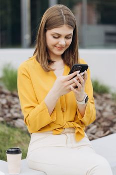 Happy businesswoman using mobile phone at remote workplace. Smiling woman browsing internet on smartphone near office. Vertical portrait of business woman reading messages on cellphone.