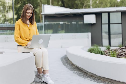 Busy worker freelancer working on modern tech notebook device. Young european business woman freelancer sitting on bench working with laptop in city park on modern urban street background.