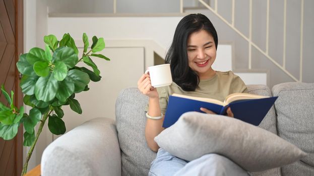 Relaxed young woman reading book and drinking hot coffee on comfortable sofa at home.