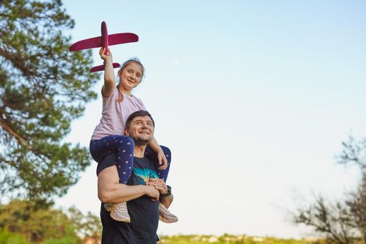 Cute girl riding on father's shoulder and playing with toy airplane against sky in summer forest