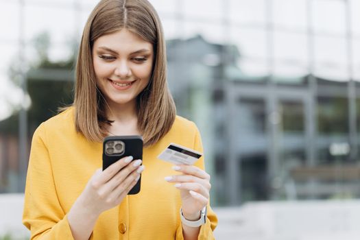Smiling girl holding credit card and smartphone sitting on bench city buildings outdoor. Happy female shopper using instant easy mobile payments making purchase in online store. E-banking app service