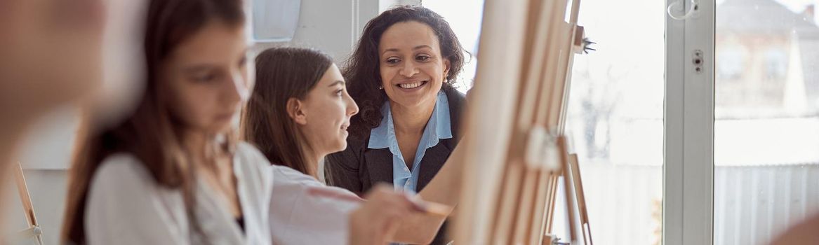 Young beautiful confident teacher is helping a kid to draw on a group lesson in a white modern minimalistic classroom.