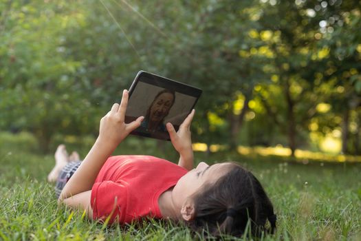 Distance learning during the quarantine period for the virus Covid-19. Cute little schoolgirl with long hair is studying from home, sitting in the garden on the grass. Uses a tablet and remote work