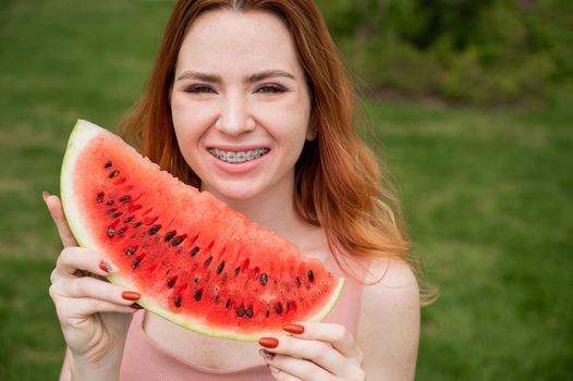 Beautiful red-haired woman smiling with braces and about to eat a slice of watermelon outdoors in summer.