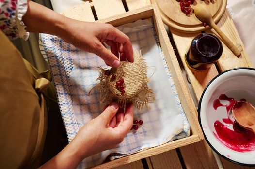 Preparing preserves, marmalades, confitures and jelly at home kitchen. Close-up hands of a housewife tying bow on the burlap on a cover, decorating a jar of homemade red currant berry jam.
