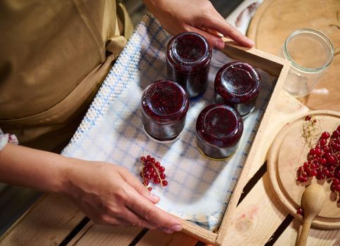 Top view of a housewife holding wooden crate with jars of berry jam stacked upside down on a waffle towel. Red currant berries and kitchen spoon on a cutting board at home rustic kitchen. Close-up