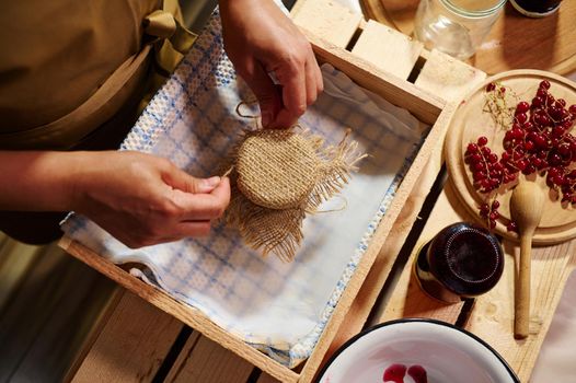 Top view of a housewife tying bow on the burlap of a lid, decorating a jar of homemade delicious jelly from red currant berries. Preparing canned food, marmalades, confitures and jams at home kitchen
