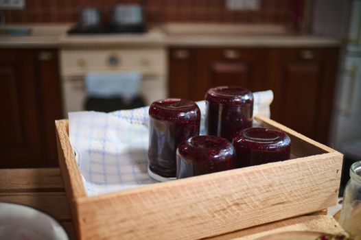 Focus on jars with berry jam stacking upside down on a wooden crate, standing on kitchen island in the home rustic cozy kitchen interior. Canned and preserved food for winter season