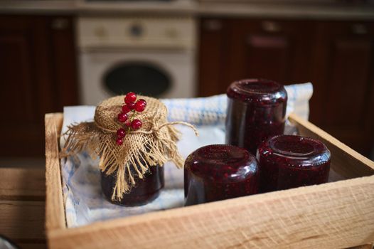 Red currant berry confitures in jars stacked upside down in a wooden box, and one bottle of sweet tasty jelly ornate with a burlap and berries on the cover in a cozy home rustic kitchen.