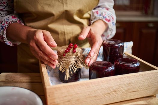 Woman hands next to a jar with jam, ornate with red currant berries on the burlap on the cover, next to jars stacked upside down on a wooden crate at home kitchen. Collection of home preserves