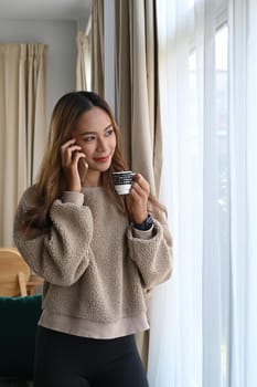 Attractive asian woman drinking coffee and talking mobile phone in living room.