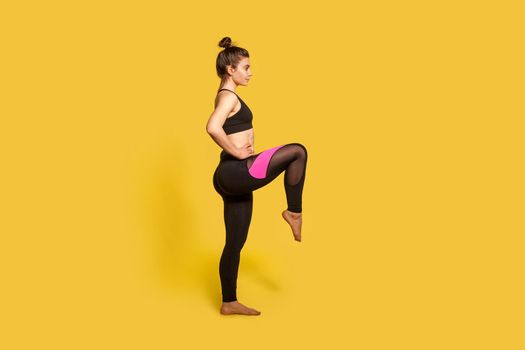 Side view, athletic slim woman with hair bun in tight sportswear doing sport standing with one leg lift, thighs workout, warming up training muscles. full length studio shot, isolated on yellow