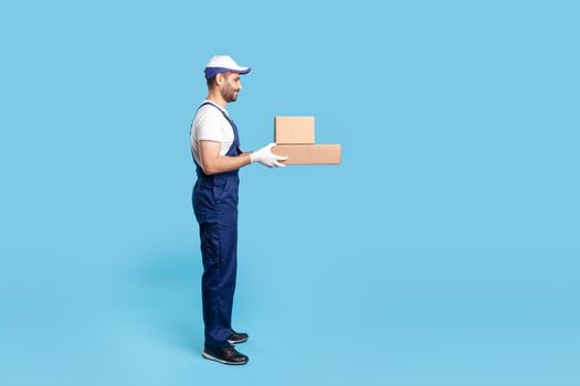 Side view expert delivery man in uniform with gloves giving cardboard boxes to client and smiling, carrying ordered purchase. Profession of courier, shipment and cargo transportation service. isolated