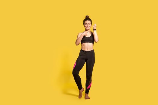 Beautiful happy athletic woman with hair bun in tight sportswear pointing at wristwatch, showing fitness tracker, electronic gadget application for sport trainings. full length studio shot, isolated