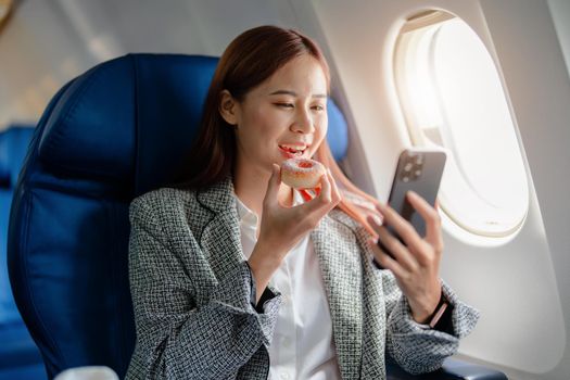 portrait of A successful asian businesswoman or female entrepreneur in formal suit in a plane sits in a business class seat and eating and using smartphone during flight.