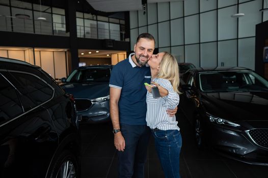 Happy caucasian couple hugging and woman holding new car keys in car showroom