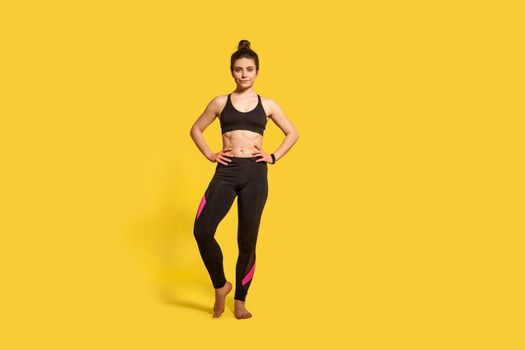 Fitness and sport training. Beautiful athletic slim sportswoman with hair bun in tight yoga pants looking at camera with confident motivated expression. studio shot, isolated on yellow background