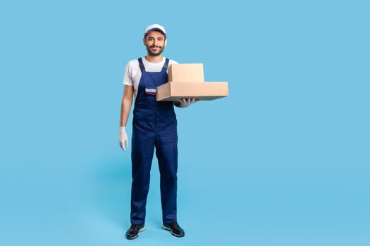 Full length delivery man in blue uniform with gloves standing, holding cardboard boxes and smiling to camera. Profession of courier, shipment and cargo transportation service. studio shot isolated