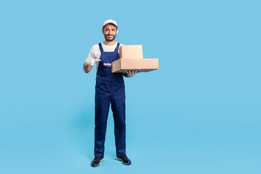 Full length cheerful delivery man in blue uniform with gloves pointing cardboard boxes and smiling to camera. Profession of courier, shipment and cargo transportation service. studio shot isolated