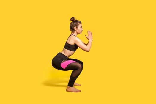 Athletic woman with hair bun in tight sportswear doing squat, lower body sport exercise, keeping balance, warming up and training muscles. full length studio shot, isolated on yellow background