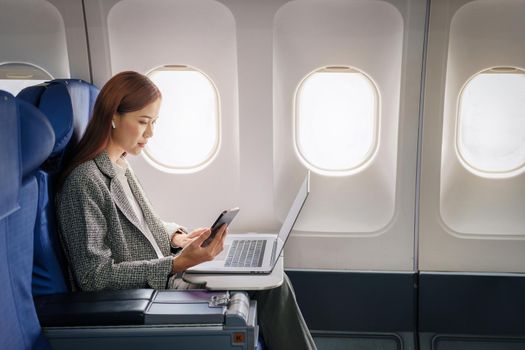 A successful asian businesswoman or female entrepreneur in formal suit in a plane sits in a business class seat and uses a documents with computer laptop during flight.