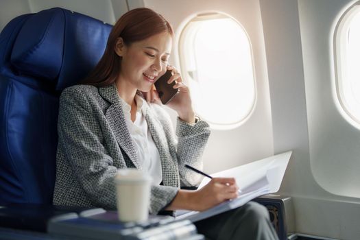 portrait of A successful asian business woman or female entrepreneur in formal suit in a plane sits in a business class seat and uses smartphone with documents for work during flight.