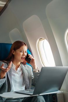 Portrait of a successful Asian businesswoman or entrepreneur in a formal suit on an airplane sitting in business class using a phone, computer laptop with a serious expression during the flight.