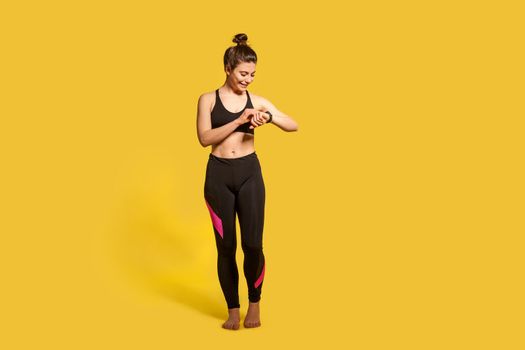 Cheerful athletic woman with hair bun in tight sportswear touching wristwatch display, checking pulse time on fitness tracker, smiling satisfied with health indicators after trainings. Sport activity