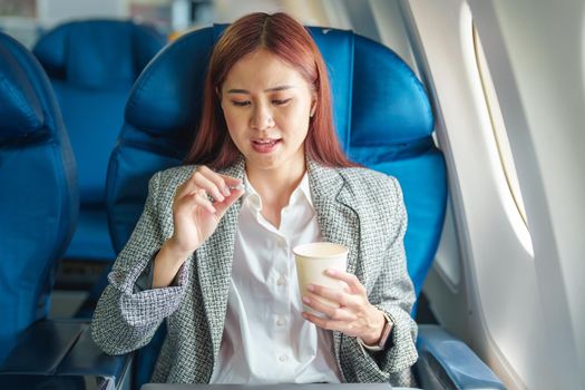 Portrait of a successful Asian businesswoman or entrepreneur in a formal suit on an airplane in business class taking motion sickness pills during the flight.