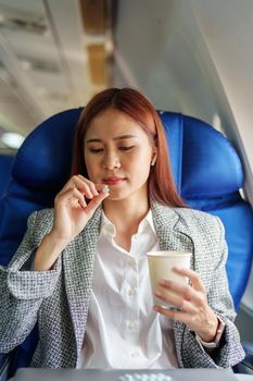 Portrait of a successful Asian businesswoman or entrepreneur in a formal suit on an airplane in business class taking motion sickness pills during the flight.