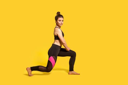 Athletic slim young woman with hair bun in tight sportswear doing sport lunge exercise, standing one knee, warming up training muscles, butts workout. full length studio shot, isolated on yellow