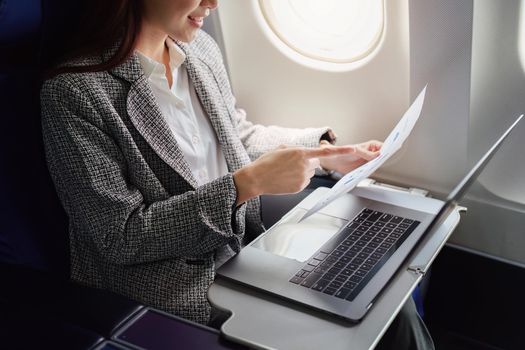A successful asian businesswoman or female entrepreneur in formal suit in a plane sits in a business class seat and uses a documents with computer laptop during flight.