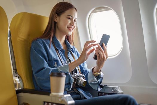 Attractive portrait of Asian woman sitting at window seat in economy class using mobile phone during plane flight, travel concept, vacation.