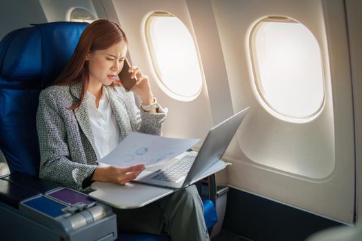 Portrait of a successful Asian businesswoman or entrepreneur in a formal suit on an airplane sitting in business class using a phone, computer laptop with a serious expression during the flight.