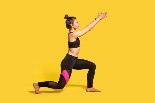 Side view, slim young woman with hair bun in tight sportswear doing sport lunge exercise, standing one knee and raising hands, warming up training muscles. full length studio shot, isolated on yellow