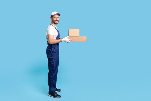 Side view friendly delivery man in uniform with gloves holding cardboard boxes and smiling to camera, carrying ordered purchase. Profession of courier, shipment and cargo transportation service