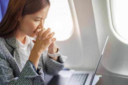 portrait of a successful Asian businesswoman or entrepreneur in a formal suit on an airplane seated in Business Class shows a thoughtful and stressed face with using laptop during the flight.