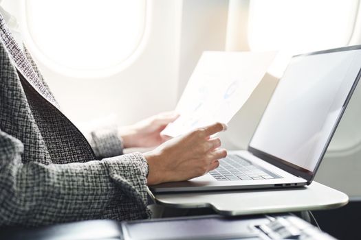 A successful asian businesswoman or female entrepreneur in formal suit in a plane sits in a business class seat and uses a documents with computer laptop during flight.