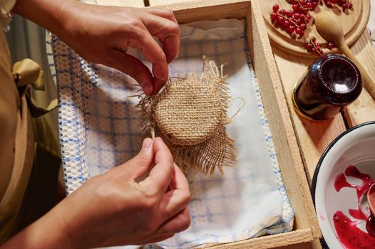 Close-up hands of a housewife tying bow on the burlap on a cover, decorating a jar of homemade red currant berry jam. Preparing preserves, marmalades, confitures and jelly at home kitchen