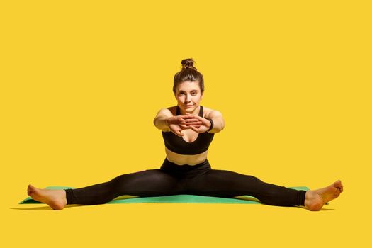 Fit gymnast woman with hair bun in tight sportswear sitting on mat with spread legs and stretching hands, warming up, doing sports flexibility exercises. studio shot isolated on yellow background