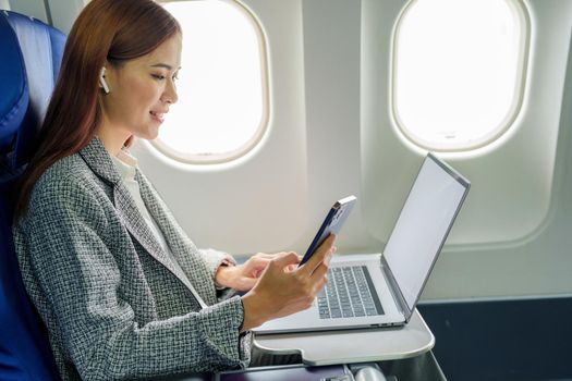 A successful asian businesswoman or female entrepreneur in formal suit in a plane sits in a business class's seat and uses a smartphone during flight.