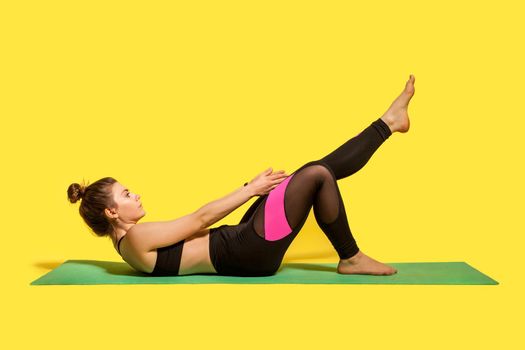 Motivated fitness woman with hair bun in yoga pants training on gym mat, reaching hands to feet stretching muscles, warming up with flexibility exercise. studio shot isolated on yellow background